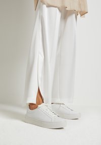 Hvite sneakers med en glatt lærfinish, flate gummisåler og minimalistisk snøring, kombinert med løse hvite bukser med sidespalter.