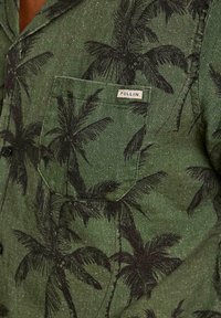 Chemise verte à boutons avec des motifs de palmiers noirs. Dotée d'une poche poitrine et d'une étiquette indiquant "PULLIN". Tissu texturé avec un léger éclat.