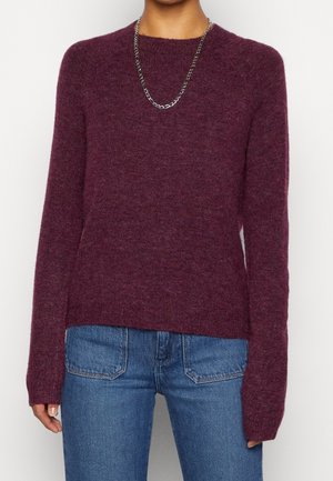 Femme portant un pull en maille bordeaux à manches longues, un jean bleu en denim avec des poches avant et un collier chaîne en argent.
