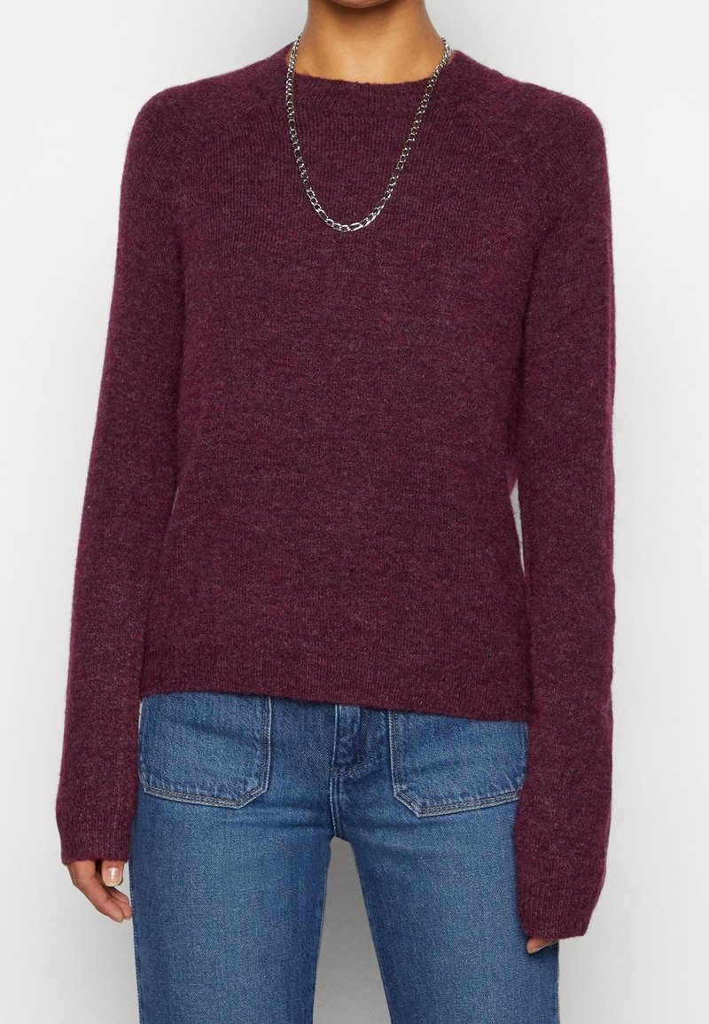 Femme portant un pull en maille bordeaux à manches longues, un jean bleu en denim avec des poches avant et un collier chaîne en argent.