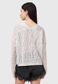 Leichtes Strickpullover mit V-Ausschnitt, offenem Strickmuster und unregelmäßigem Textdesign in einem hellgrauen Farbton, kombiniert mit dunklen Shorts.