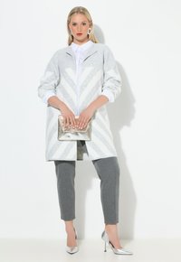 Grijze en witte gestreepte lange cardigan, grijze slim-fit broek, zilveren hoge hakken, met een metallic clutch in de hand, en daaronder een witte blouse met kraag.