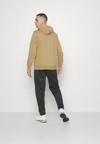 Felpa con cappuccio color tan, dotata di polsini e orlo a costine, abbinata a joggers neri a gamba stretta. Il modello indossa sneakers bianche. Sfondo semplice.