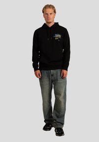 Felpa nera con un design grafico caratterizzato da accenti blu e gialli. Abbinata a jeans azzurri chiari e sneakers nere. Silhouette dal fit rilassato.