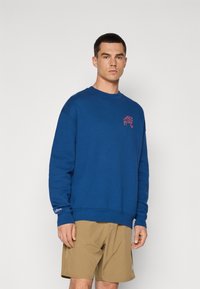 Sweatshirt azul feito de algodão, com gola redonda, mangas longas e um pequeno detalhe bordado. Combinado com calções caqui.