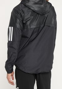 Czarna, lekka kurtka z kapturem, o błyszczącej teksturze i kontrastowej matowej sekcji. Trzy białe paski na lewym rękawie. Regulowane mankiety.