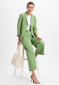 Grüne Blazer und passende weite Hosen, kombiniert mit einem weißen Oberteil, einer beigen Handtasche und braunen Riemchenschuhen. Glatter Stoff und taillierte Passform.