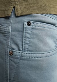 Pantalones de mezclilla de color azul claro que cuentan con un bolsillo frontal con un botón a presión negro, detalles de costura y una prenda texturizada verde por encima.