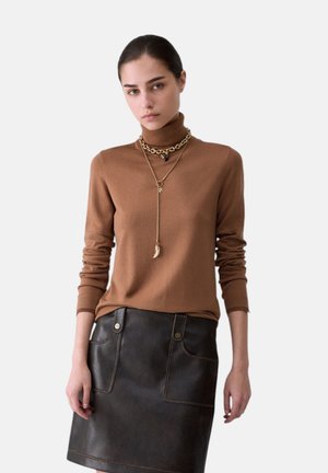 Donna che indossa un maglione marrone a collo alto e una gonna nera in pelle con tasche frontali, accessoriata con collane a catena dorate a strati.