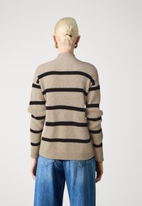 Beżowy sweter w prążki z czarnymi poziomymi paskami, wysokim kołnierzem i długimi rękawami, zestawiony z niebieskimi szerokimi spodniami typu jeans.