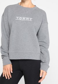 Vrouw in een grijze sweatshirt met lange mouwen met het tekst "TOMMY" op de borst en een zwarte legging, staand met ontspannen handen langs de zij.