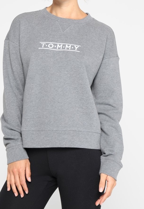 Vrouw in een grijze sweatshirt met lange mouwen met het tekst "TOMMY" op de borst en een zwarte legging, staand met ontspannen handen langs de zij.