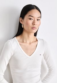 White ribbed long-sleeve top with a deep V-neck, featuring a small logo on the chest. Model wears large hoop earrings. Smooth texture.