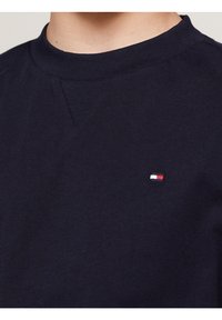 Námořnická modrá bavlněná tričko s žebrovaným kulatým výstřihem a malým vyšitým logem v červené, bílé a modré barvě na levé části hrudi.