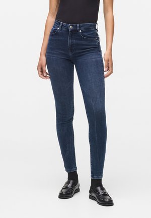 Femme portant un jean skinny bleu foncé, un haut noir ajusté, des chaussettes noires et des mocassins noirs cloutés, debout devant un fond blanc uni.