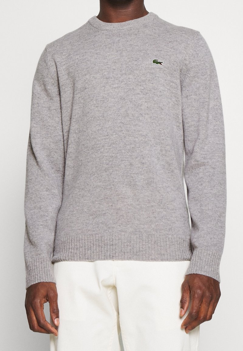Homme portant un pull gris clair à col rond avec un petit logo crocodile vert sur la poitrine et un pantalon blanc, debout devant un fond uni.