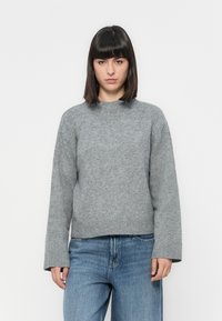 Donna con capelli neri lisci, che indossa un maglione grigio a maglia e jeans blu, in piedi davanti a uno sfondo bianco semplice.