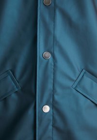 Jachetă impermeabilă de culoare teal, cu o textură netedă, având nasturi metalici argintii și două buzunare laterale. Cusături curate de-a lungul placket-ului.