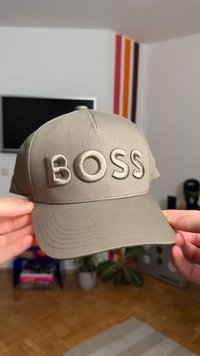 Pelēka cepure ar izliekta forma briļļu aizsegu, uz kura ir izcelts "BOSS" logotips pelēkā krāsā. Izgatavota no gluda, mīksta materiāla. Redzamas dekoratīvās šuves.
