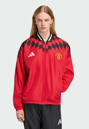 Ung person med langt hår iført en rød Manchester United jakke med sort diamantmønster og sorte Adidas bukser mod en ensfarvet baggrund.