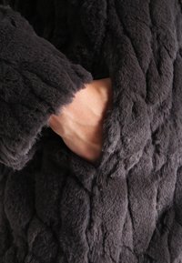 Grijze nepbontjas met een gewatteerde structuur, voorzien van een zijzak en zacht, pluizig materiaal, dat het dikke en comfortabele ontwerp benadrukt.