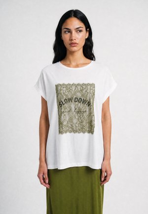Junge Frau mit langen dunklen Haaren trägt ein weißes T-Shirt mit grünem Spitzenquadrat und "Slow down and loose" Text über einem olivgrünen Rock.