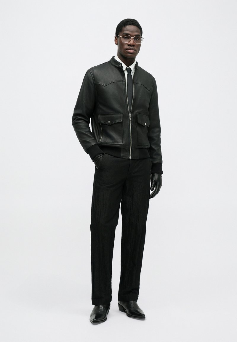 Black leather jacket with two front pockets, paired with textured black trousers and black leather shoes. Model wears thin glasses and gloves.