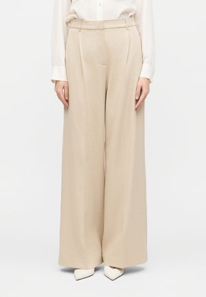 Béžové široké nohavice s vysokým pásom spojené s bielymi topánkami so špicatou špičkou a bielou dlhorukávovou košeľou, zobrazené od pása nadol.