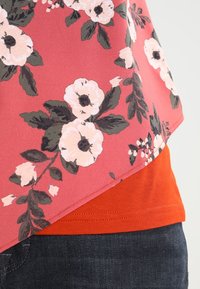 Top floreale con sfondo corallo, con grandi fiori bianchi e foglie verdi. Sotto, è visibile una maglietta rossa a tinta unita, abbinata a jeans scuri.