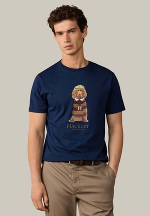 Joven con cabello rizado que lleva una camiseta azul marino con la imagen de un perro con suéter a rayas y pantalones beige con un cinturón trenzado.