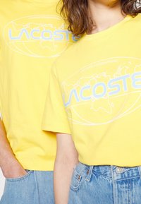 Žluté bavlněné trička s kulatým výstřihem, na kterých je modrá grafika světové mapy a nápis "LACOSTE INTERNATIONAL". Volný střih, krátké rukávy.