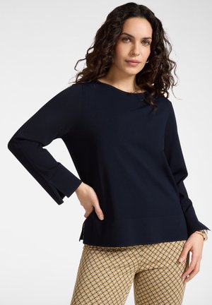 Blusa blu navy a maniche lunghe con scollo tondo, polsini piegati e taglio dritto. Abbinata a pantaloni beige e gialli con fantasia.