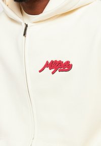 Crèmekleurige zip-up hoodie met een rood geborduurd logo. Heeft een gladde stoftextuur en een capuchon, met een minimalistisch ontwerp.