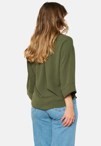 Olive green, loose-fitting top with three-quarter sleeves and a round neckline, paired with light blue jeans. Soft texture, casual design.