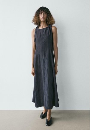 Donna con capelli ricci che indossa un vestito senza maniche grigio scuro fino alla caviglia e scarpe nere, in piedi contro un muro chiaro e semplice.