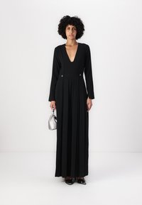 Schwarzeslangärmliges Kleid mit tiefem V-Ausschnitt, plissiertem Rock und Knopfakzenten an der Taille. Model hält eine silberne Tasche und trägt schwarze High Heels.