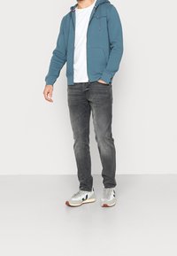 Sudadera con cremallera de color azul claro, con bolsillos, llevada sobre una camiseta blanca, combinada con jeans gris oscuro y zapatillas grises con acentos en azul marino.