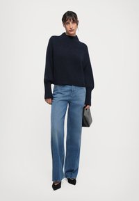 Marineblauwe gebreide trui met hoge kraag, gecombineerd met lichtblauwe wijde jeans. Een zwarte hoekige handtas en puntige zwarte hakken maken de look compleet.