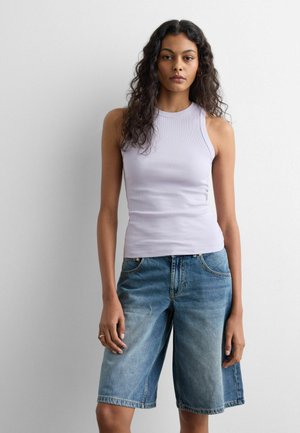 Junge Frau mit lockigem Haar trägt ein ärmelloses lavendelfarbenes Top und kniehohe Jeansshorts, steht vor einem einfachen weißen Hintergrund.