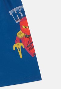 Plava majica s printanim crvenim ninja likom i žutom oružjem. Dizajn uključuje odvažne grafike i glatku teksturu tkanine.