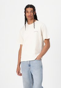 Cream-colored cotton t-shirt with a relaxed fit, featuring a small, curved logo text in brown on the left chest. Paired with light blue jeans.
