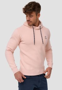 Svijetlopinkast dukserica s kapuljačom koja se može stegnuti, rebrastim manžetama i donjim rubom, koja na prsima ima mali izvezeni logotip i mekani, strukturirani materijal.