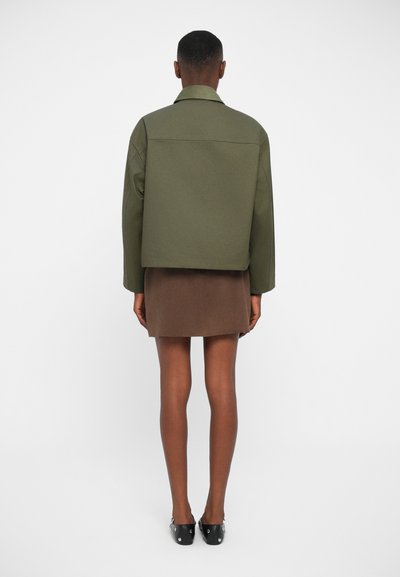 Olive green cropped jacket with a simple design and structured collar, paired with a brown skirt. Model stands with back to camera.