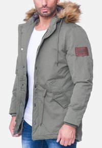 Parka verde oliva con cappuccio bordato di pelliccia, bottoni a scatto frontali, tasche laterali e una toppa di pelle sulla manica. Interno foderato con motivo a quadrato.