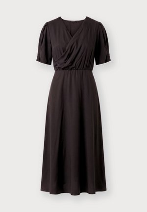 Femme debout face à l’avant portant une robe midi noire à manches bouffantes et décolleté en V drapé, assortie à des chaussures noires à bout pointu.