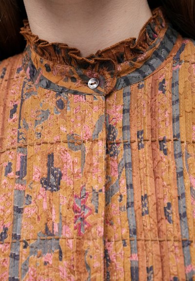 Aukšta apykaklė su raukiniais, užsegama mygtukais, su gėlių raštu garstyčių, rožinės, pilkos ir mėlynos vertikalios juostos.