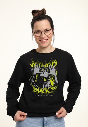 Frau mit Brille und Dutt lächelt und trägt einen schwarzen Sweatshirt mit einer Grafik eines Hundes, der die Zunge herausstreckt, und Text in Gelb und Weiß.