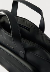 Melnā ādas soma ar fakturētu apdari, divām rokturu siksnām, rāvējslēdzēja aizdari un logotipa uzlīmi iekšpusē. Aprīkota ar gludu iekšējo oderi.