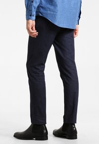 Man som bär mörkblå slim-fit byxor, svarta läder Chelsea-boots, och en blå skjorta med knappar, står avslappnat med en hand.