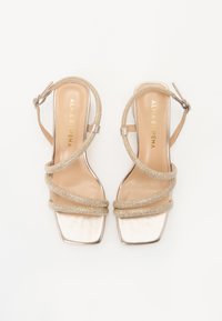 Sandales à talons hauts dorées avec des brides croisées et un embout carré. Brides en texture semblable à du mesh et boucle à l'arrière. Fini métallique lisse sur la semelle. Nom de la marque embossé à l'intérieur.